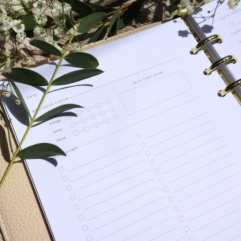 Planner with a self-care plan section on a surface with green leaves and flowers.