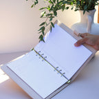 Open planner with lined pages held by a hand, with a plant in the background